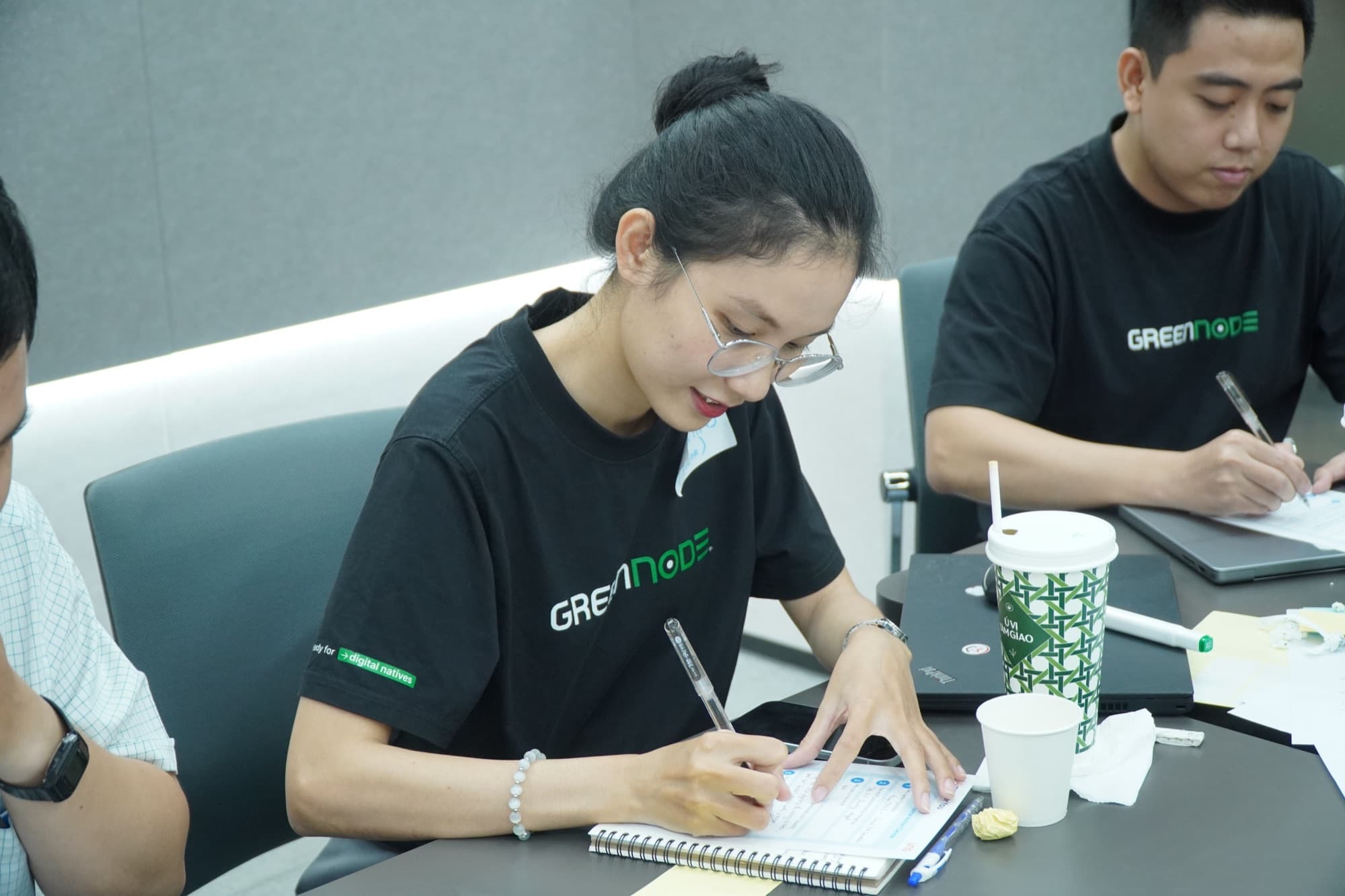 Hana ngồi làm bài tập cá nhân tại workshop, áo GreenNode, sổ tay và canvas trên bàn